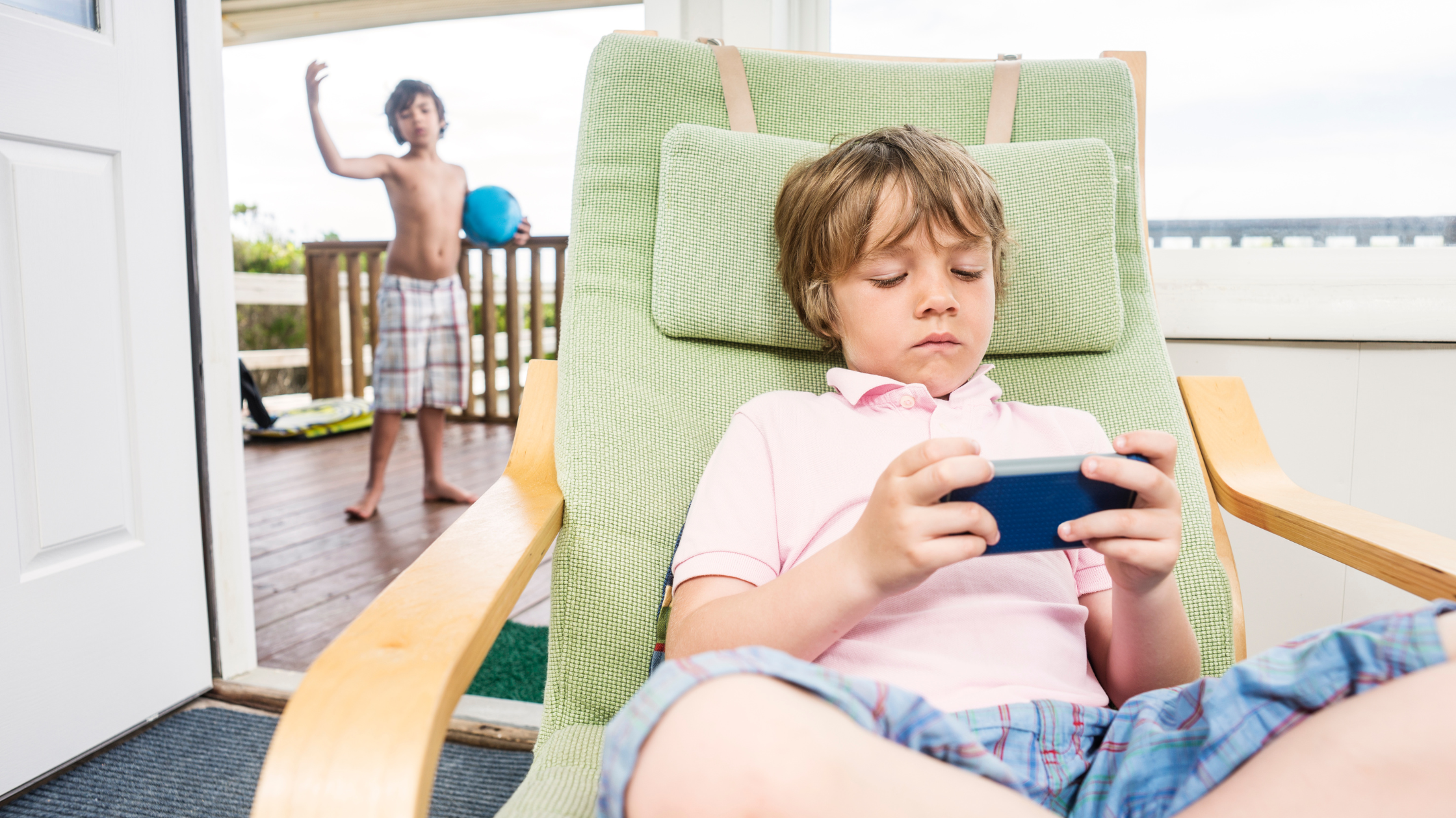 A child using a cellphone while another child in the background invites him to play ball, illustrating the potential link between screen time, reduced physical activity, and childhood obesity.