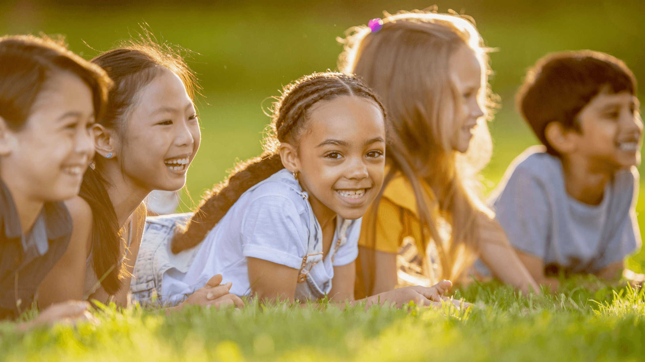 A diverse group of smiling children lying on green grass, possibly suggesting the joy of outdoor summer activities that contribute to overall well-being and healthy digestion in kids.