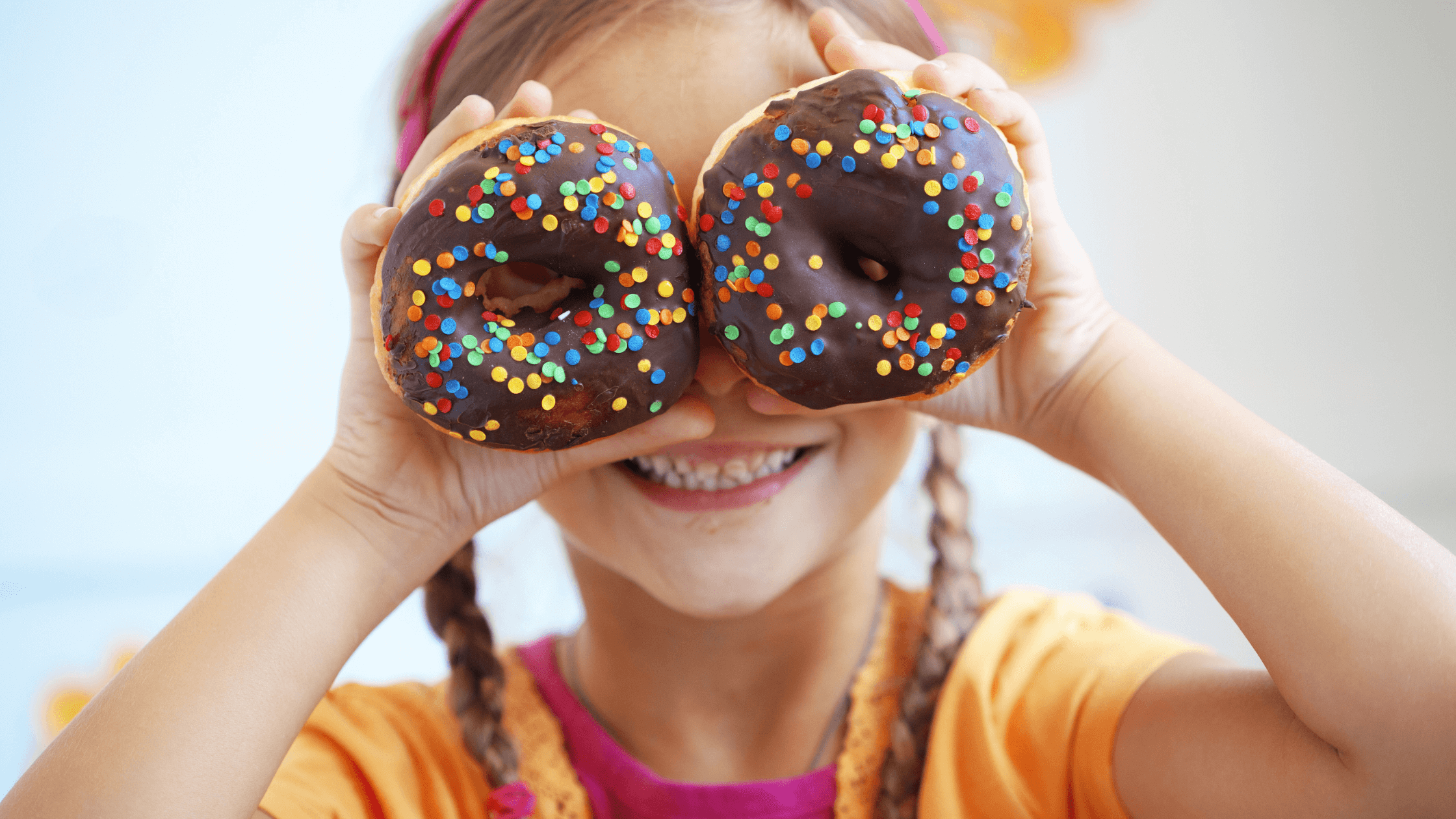 A smiling child playfully holding two chocolate-frosted donuts with colorful sprinkles over their eyes
